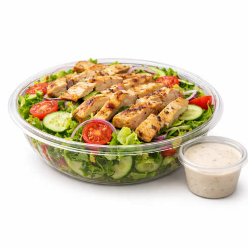Grilled chicken salad in a clear plastic takeout bowl with romaine, cucumbers, cherry tomatoes, and red onion, served with a small cup of creamy dressing, photographed on a clean white background.
